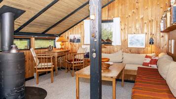 Acogedor interior de cabaña con paredes de madera, un sofá, una mesa, sillas, chimenea y ventana con vistas a la vegetación.