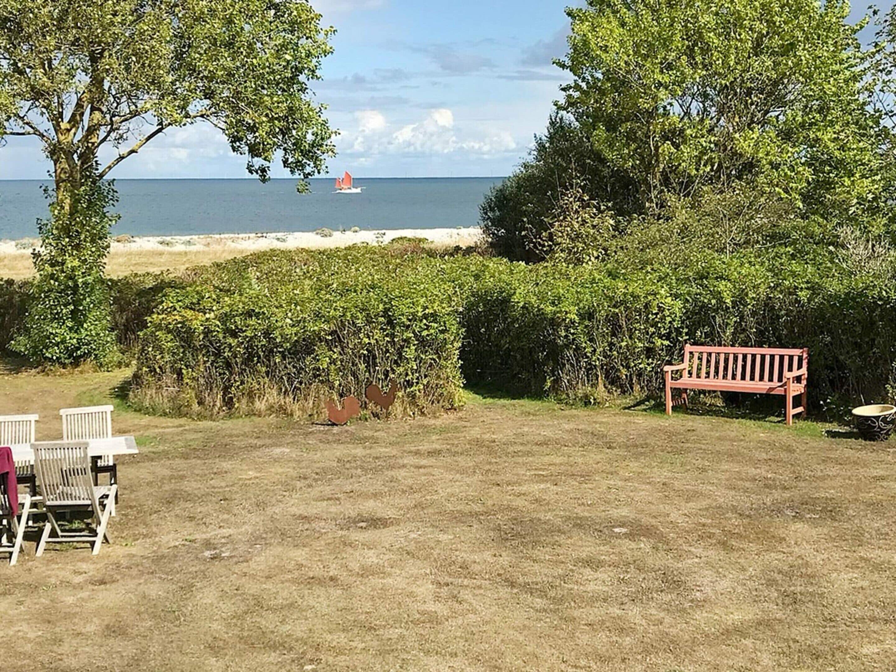 Un espacio al aire libre con césped, una banca, sillas y vista al mar con un velero.