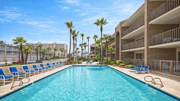 A swimming pool with blue lounge chairs, palm trees, and a multi-story building with balconies. - Padre Oasis #302 2 Bedroom Home by RedAwning (South Padre Island)