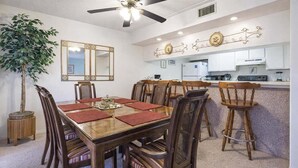 A dining area with a wooden table and chairs, a ceiling fan, a potted plant, and a kitchen area with white cabinets. - Padre Oasis #302 2 Bedroom Home by RedAwning (South Padre Island)