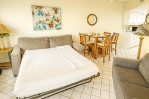 A room with a bed, a sofa, a dining table with chairs, a kitchen area, and a painting on the wall.