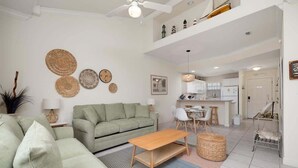 A spacious living area with a sofa, a wooden coffee table, and a dining table with chairs. There are decorative wall hangings and a kitchen area in the background.