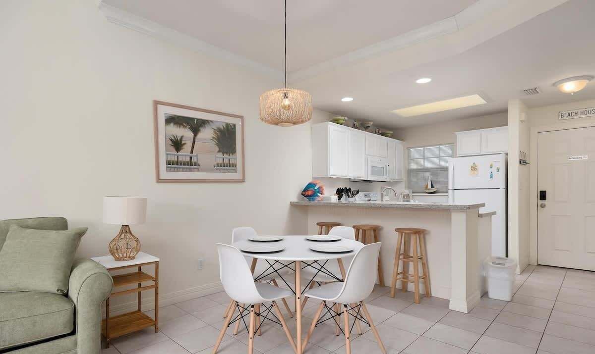 Uma cozinha moderna com armários brancos, uma área de refeições com cadeiras brancas, um lustre e um quadro na parede.