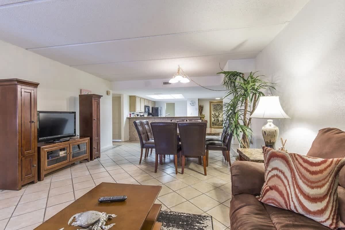 A living room with a television, a coffee table, and a sofa. There is a dining area with chairs and a table in the background.