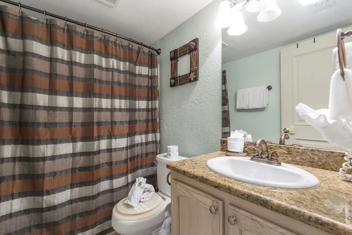 Ein Badezimmer mit gestreiftem Duschvorhang, einem Waschbecken mit Granitplatte, einer Toilette und einem Spiegel mit einem Wandtuch.