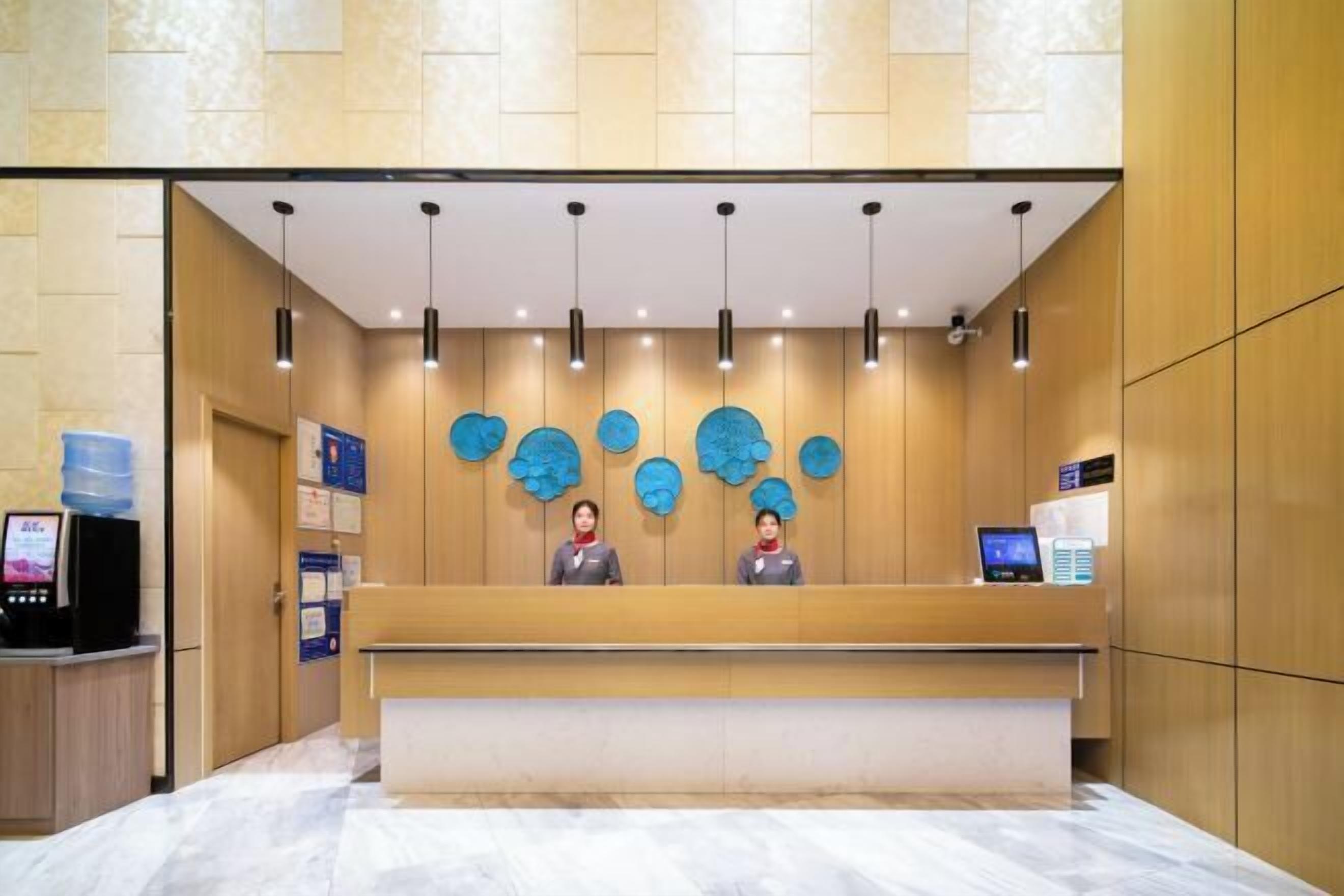 Ein Hotel-Rezeptionsbereich mit einem Marmor-Empfangstresen, zwei Mitarbeitern an der Rezeption, einem Wasserspender und einer digitalen Informationsstelle.