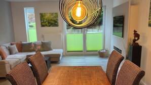 Un espace repas avec une table en bois et des chaises en osier, un luminaire suspendu et une vue sur l’extérieur grâce à de grandes fenêtres.