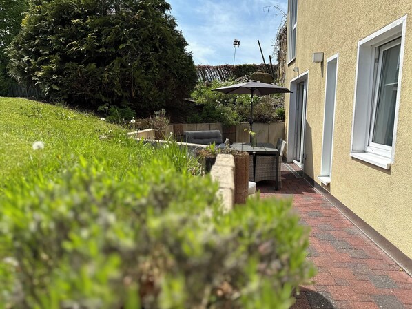 Un espace patio aménagé avec une table et des chaises, un pot de fleurs et une vue sur une haie et des arbres.