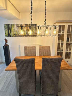 Un coin repas avec une table en bois et des chaises en osier, un luminaire suspendu avec des ampoules apparentes, et un meuble avec des portes en verre en arrière-plan.