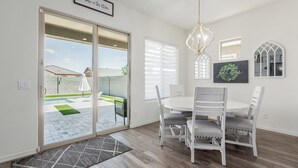 Un comedor con una mesa redonda y seis sillas, una araña y un ventanal corredero que da a un patio con piscina.