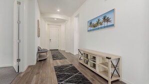 Un pasillo con piso de madera, un banco, una puerta y un cuadro de palmeras.