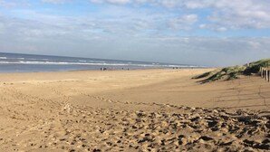 A wide sandy beach with gentle waves, a clear sky, and a grassy dune area. - Holiday Home Dune Nest (Noordwijk)