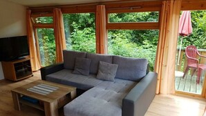 En moderne stue med en grå hjørnesofa, et træsofabord og et tv på et træstel. Store vinduer giver udsigt til grøn natur udenfor.