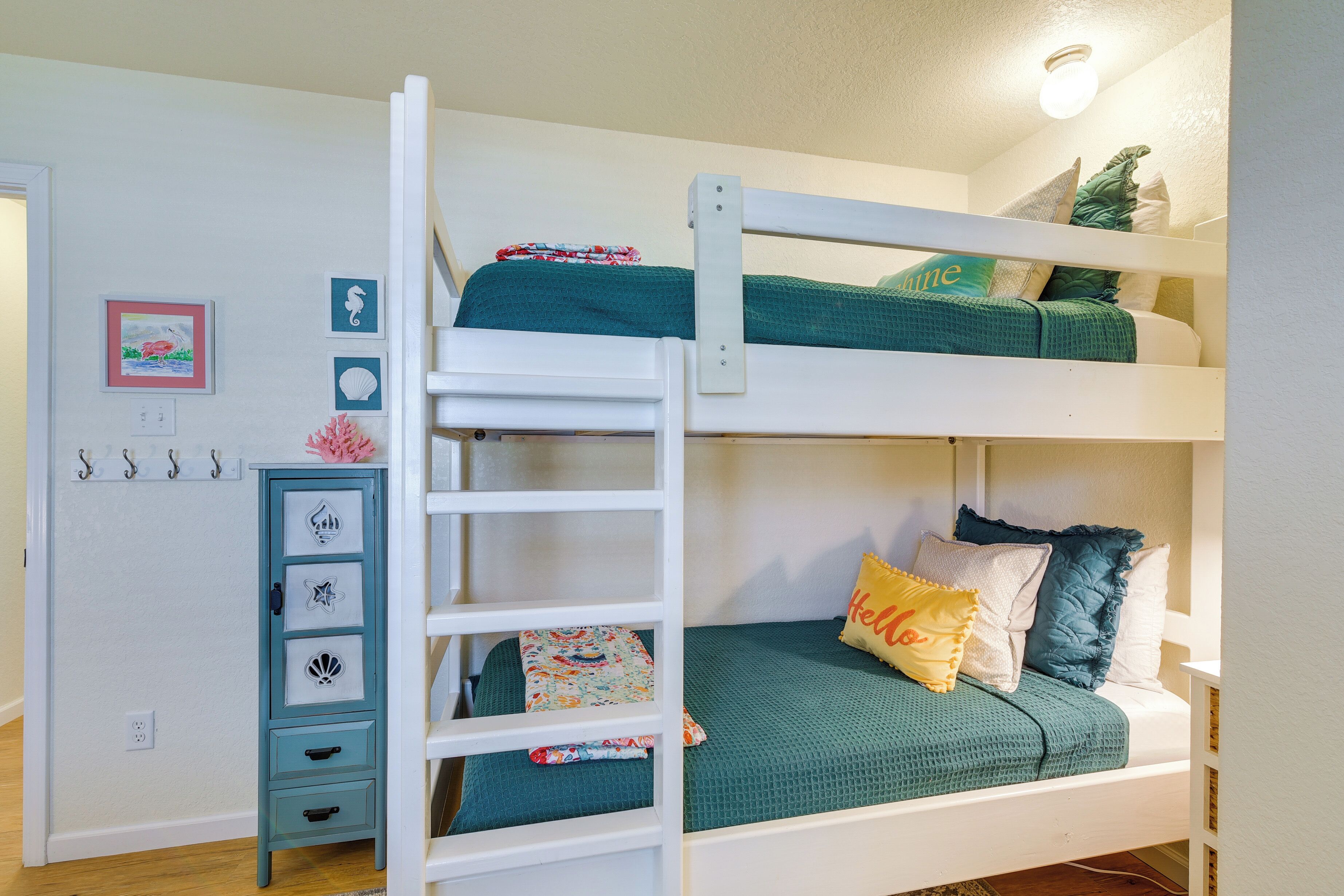 Una cama litera con escalera, un archivador pequeño y cajones, y una pared con cuadros y ganchos.