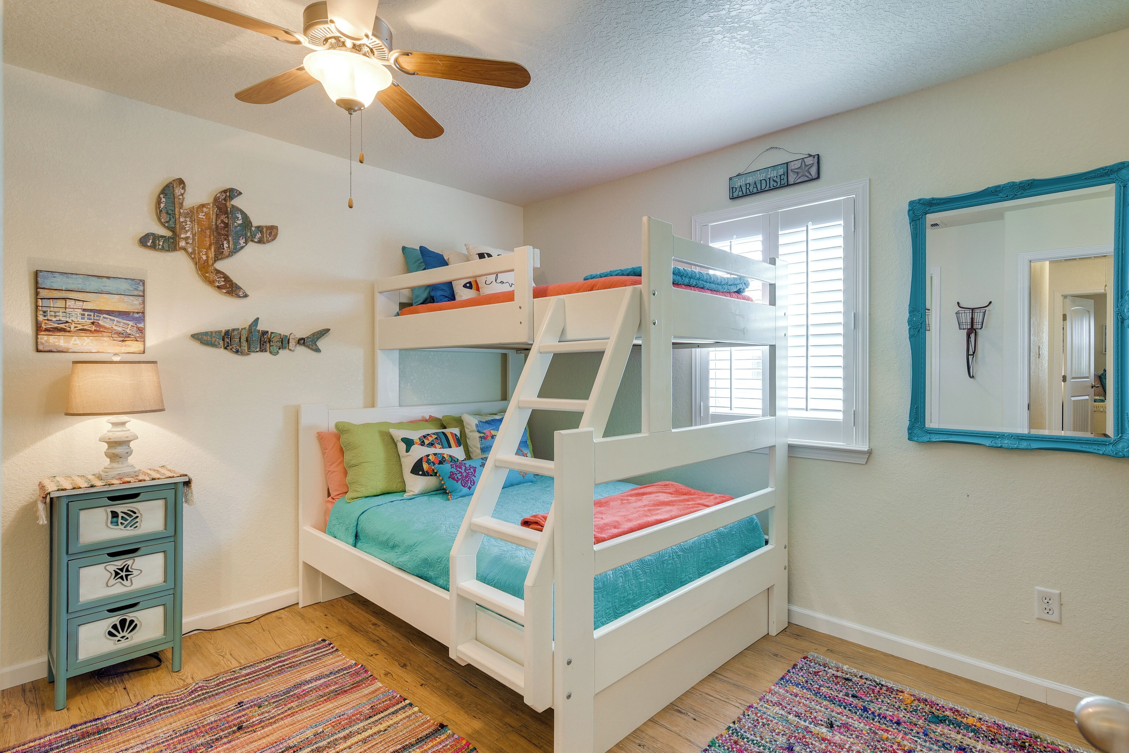 A bunk bed room with a ceiling fan, a small table with drawers, a colorful rug, and a wall with a mirror and artwork.