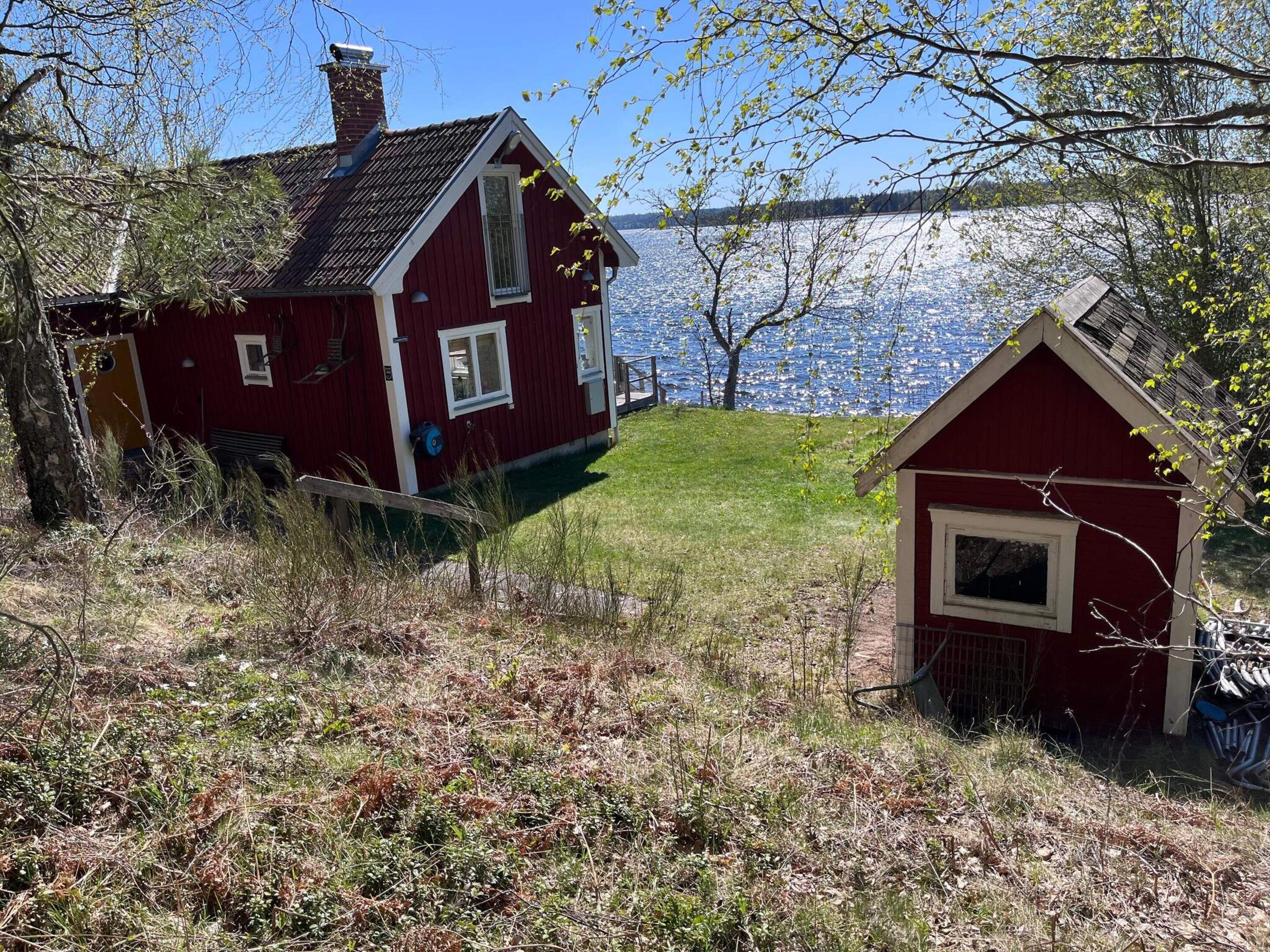 Una casa roja con chimenea, una construcción roja más pequeña y un cuerpo de agua al fondo.