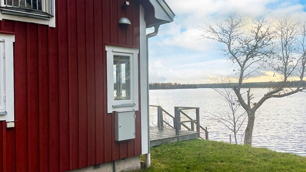 A red building with a dock extending into a lake, surrounded by trees and a clear sky. - 5 Star Holiday Home in Hjaltevad (Hjältevad)