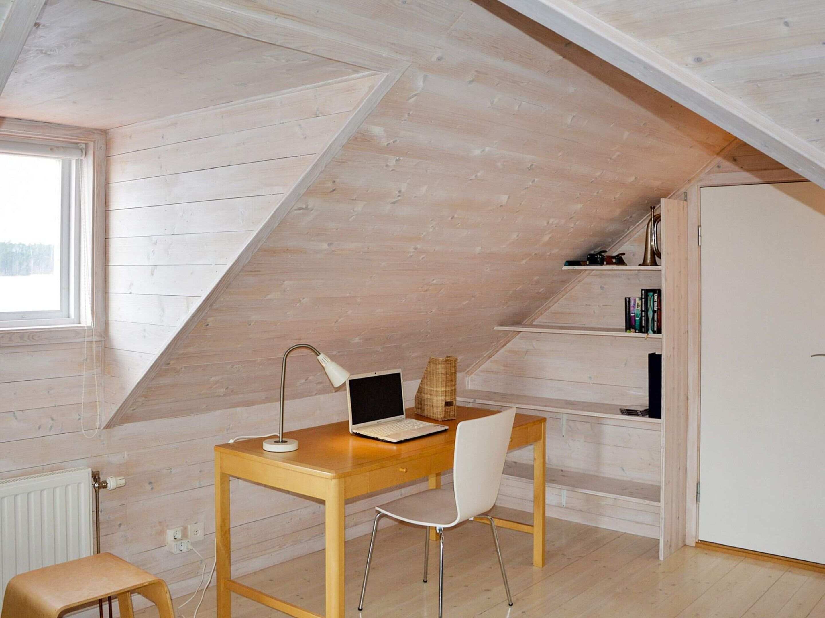 Une pièce avec un plafond en pente, un bureau en bois avec un ordinateur portable, une chaise, un radiateur et une bibliothèque.