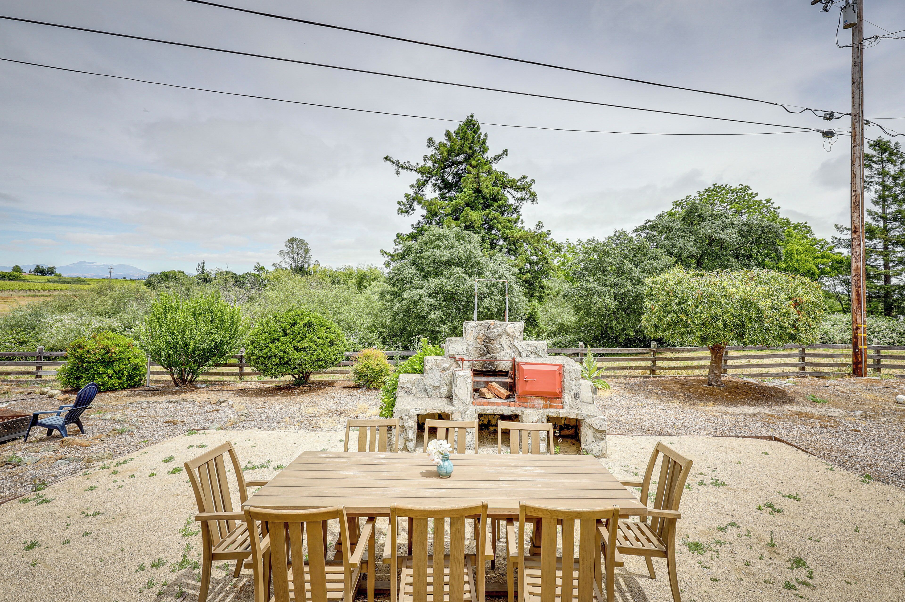 An outdoor dining area with a stone fireplace, surrounded by a gravel patio and a wooden fence.