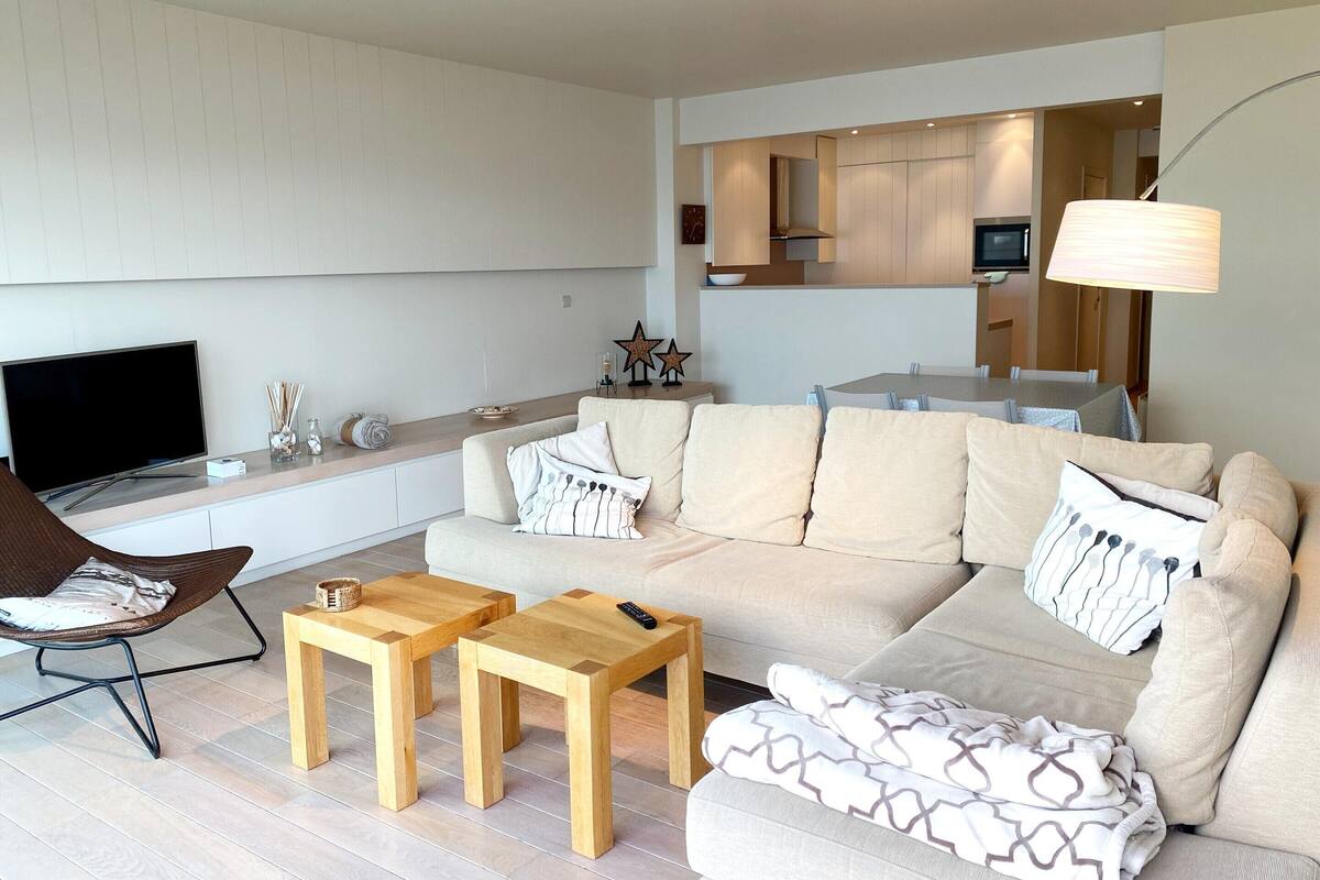 Un salon moderne avec un canapé, deux petites tables en bois, un téléviseur et, au fond, un coin repas.