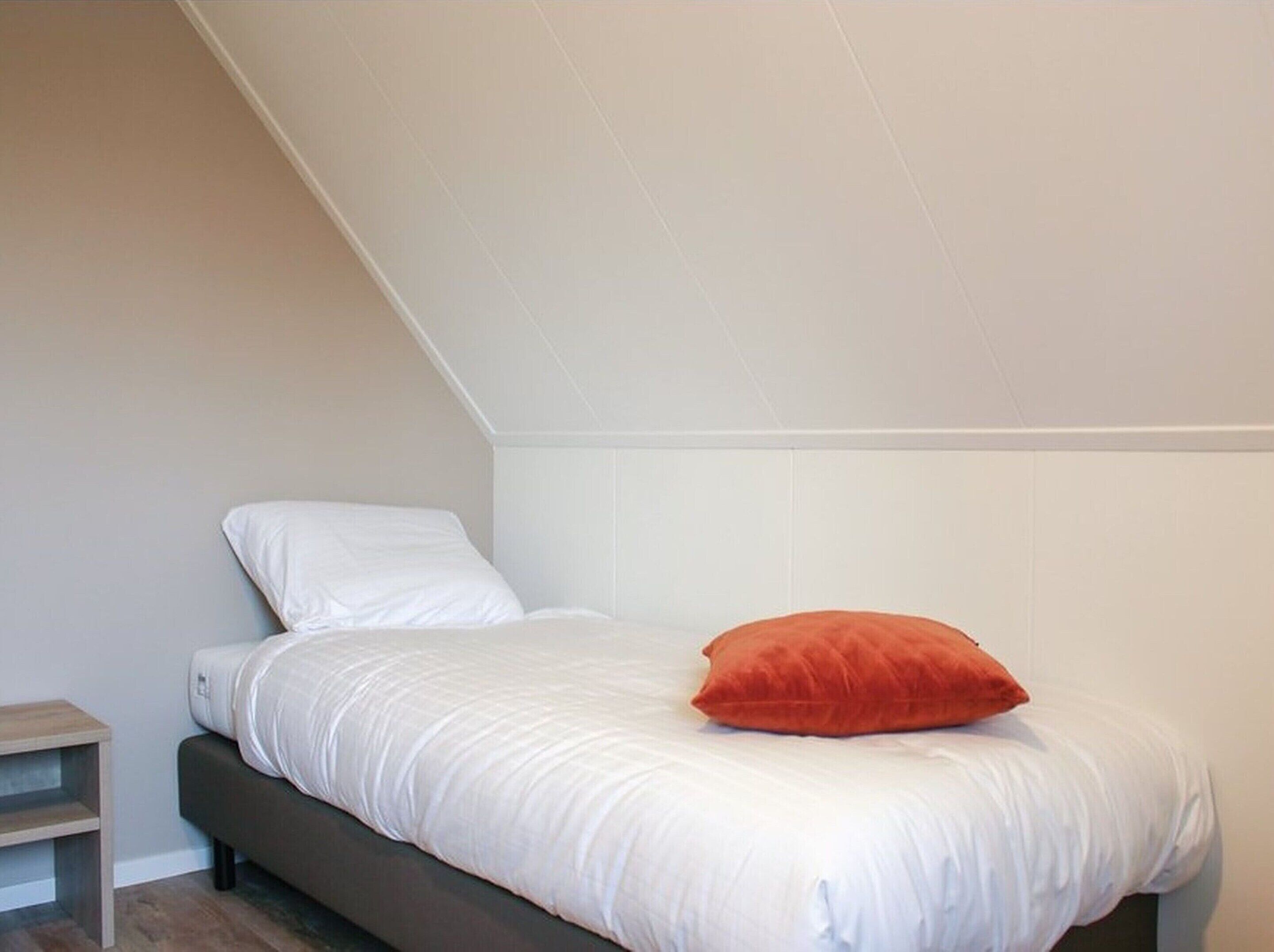 A single bed with white bedding and a red pillow, a wooden nightstand, and a sloped ceiling.