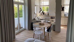 A modern living room with a sofa, dining table, and chairs, large windows, and a view of greenery outside. - Luxury & Design With sea View (Kuehlungsborn)
