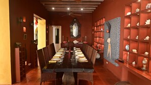 A dining room with a long table set for a meal, surrounded by chairs, with a ceiling fan above and a patterned wall to the right.