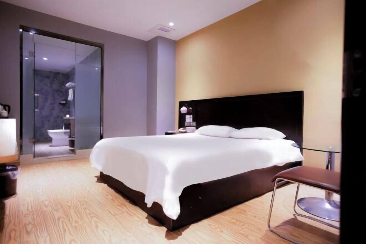 A modern hotel room with a large bed, a glass door leading to a bathroom, and a wooden floor.
