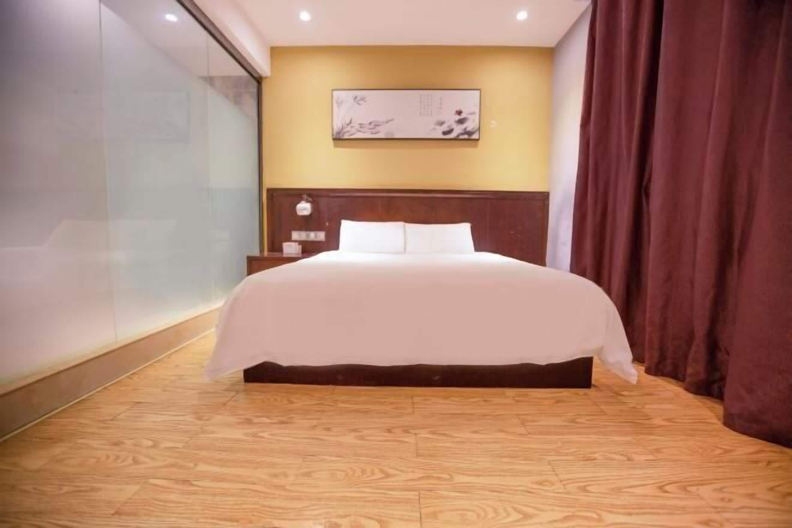 A hotel room with a large bed, wooden flooring, a nightstand, a wall-mounted artwork, and a curtain.