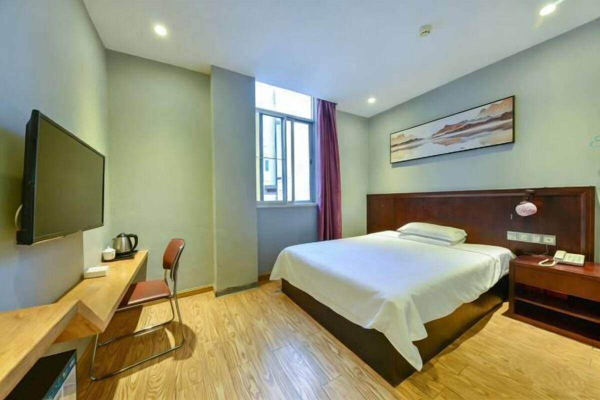 A hotel room with a bed, a desk with a chair, a TV, a window with curtains, and a painting on the wall.
