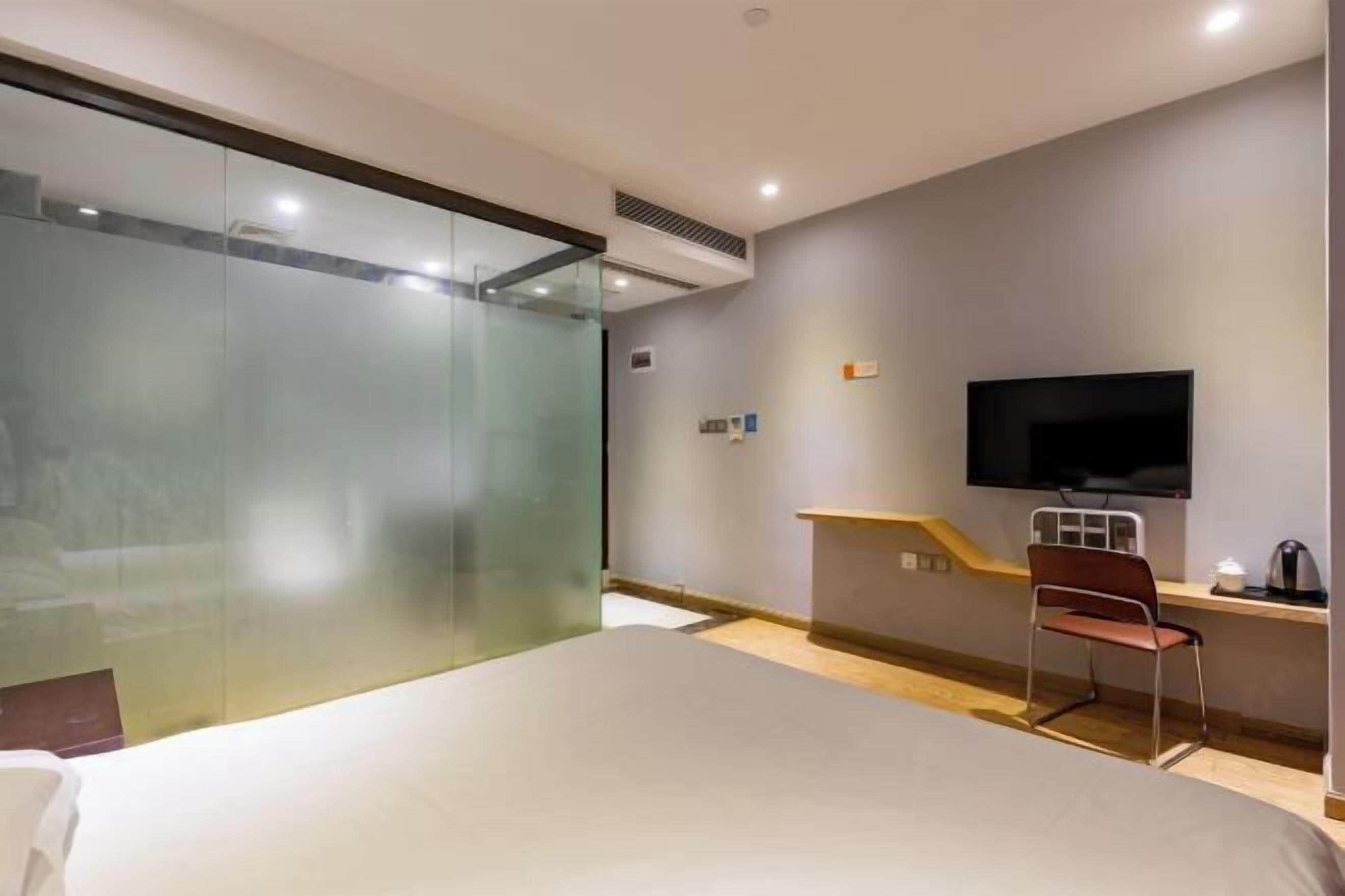 A hotel room with a bed, a desk with a chair, a television, and a glass partition.