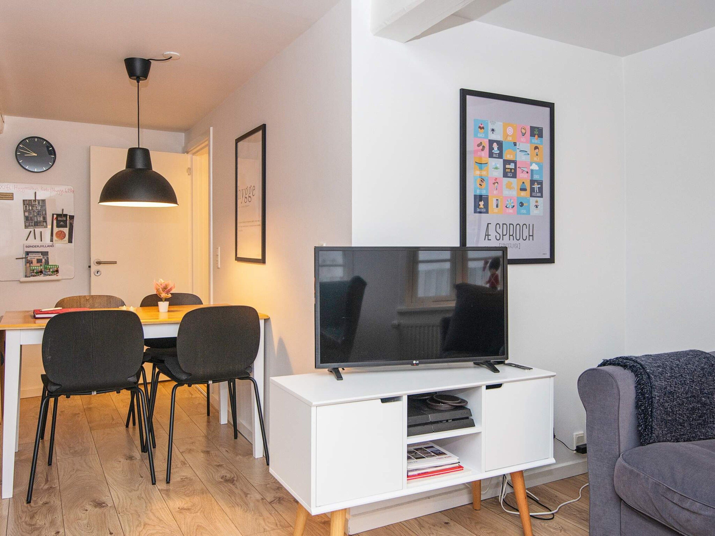 Ein modernes Wohnzimmer mit einem Flachbildfernseher auf einem weißen Schrank, einem grauen Sofa und einem Essbereich mit Tisch und Stühlen.