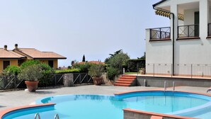 Ein Schwimmbecken mit gewölbter Kante, umgeben von Topfpflanzen und einem weißen Gebäude mit Balkon.