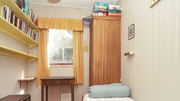 A bedroom with a wooden wardrobe, a bedside table, a bed with a blue pillow, a window with curtains, a bookshelf, and a radiator. - 5 Person Holiday Home in Rorvig (Rørvig)