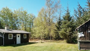 A small, single-story cabin with a white door and a chimney, surrounded by a grassy yard and dense trees. - 5 Person Holiday Home in Rorvig (Rørvig)