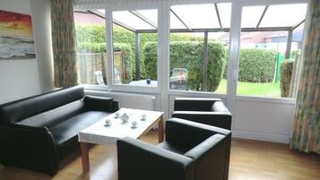 A living room with a sofa, two armchairs, and a table with teacups. Large windows offer a view of a garden and a house.