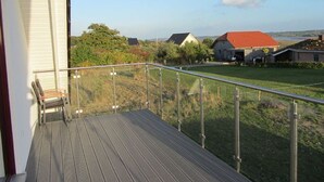 Ein Balkon mit Holzdeck und Metallgeländer mit Blick auf eine Rasenfläche, im Hintergrund Häuser.