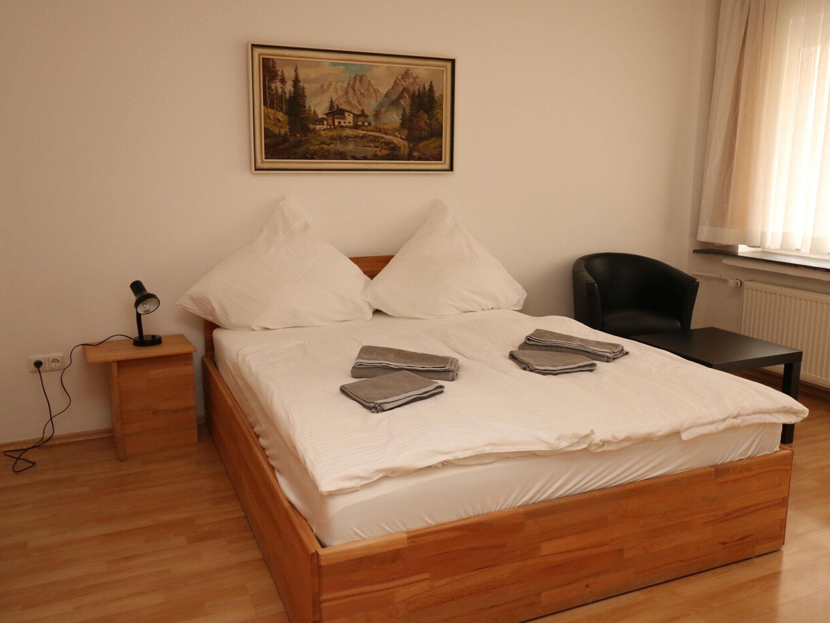 A neatly made bed with white linens, a painting of a mountain landscape on the wall, and a chair to the right.