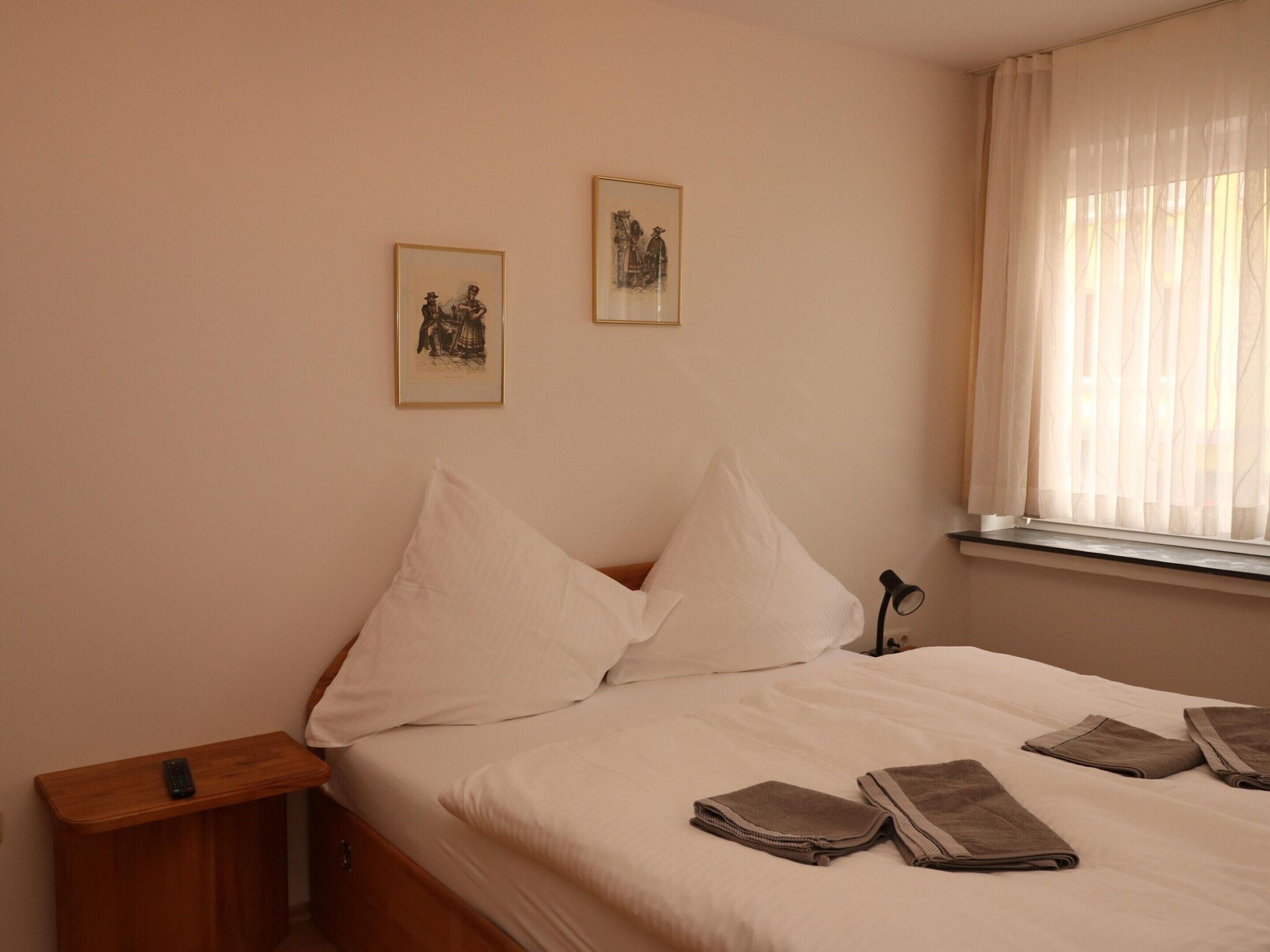 A neatly made bed with white linens, a bedside table with a remote control, and two framed pictures on the wall.