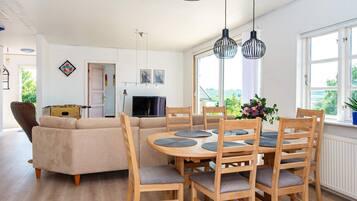 Uma sala de estar moderna com sofá, mesa de jantar e cadeiras. Há uma televisão e uma janela com vista.
