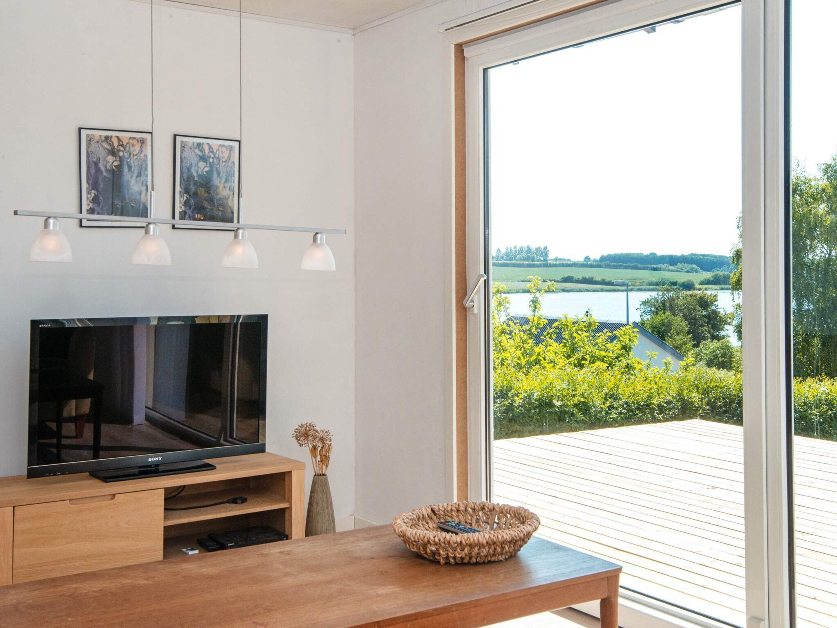 Ein modernes Wohnzimmer mit einem Flachbildfernseher, Holzmöbeln und Seeblick durch eine Schiebetür.