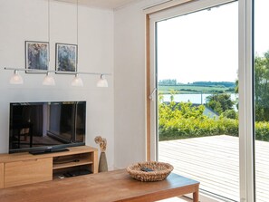 A modern living room with a flat-screen TV, wooden furniture, and a view of a lake through a sliding glass door. - 6 Person Holiday Home in Nordborg (Nordborg)