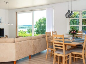 A modern living room with a sofa, dining table, chairs, and large windows offering a view of greenery and a body of water. - 6 Person Holiday Home in Nordborg (Nordborg)
