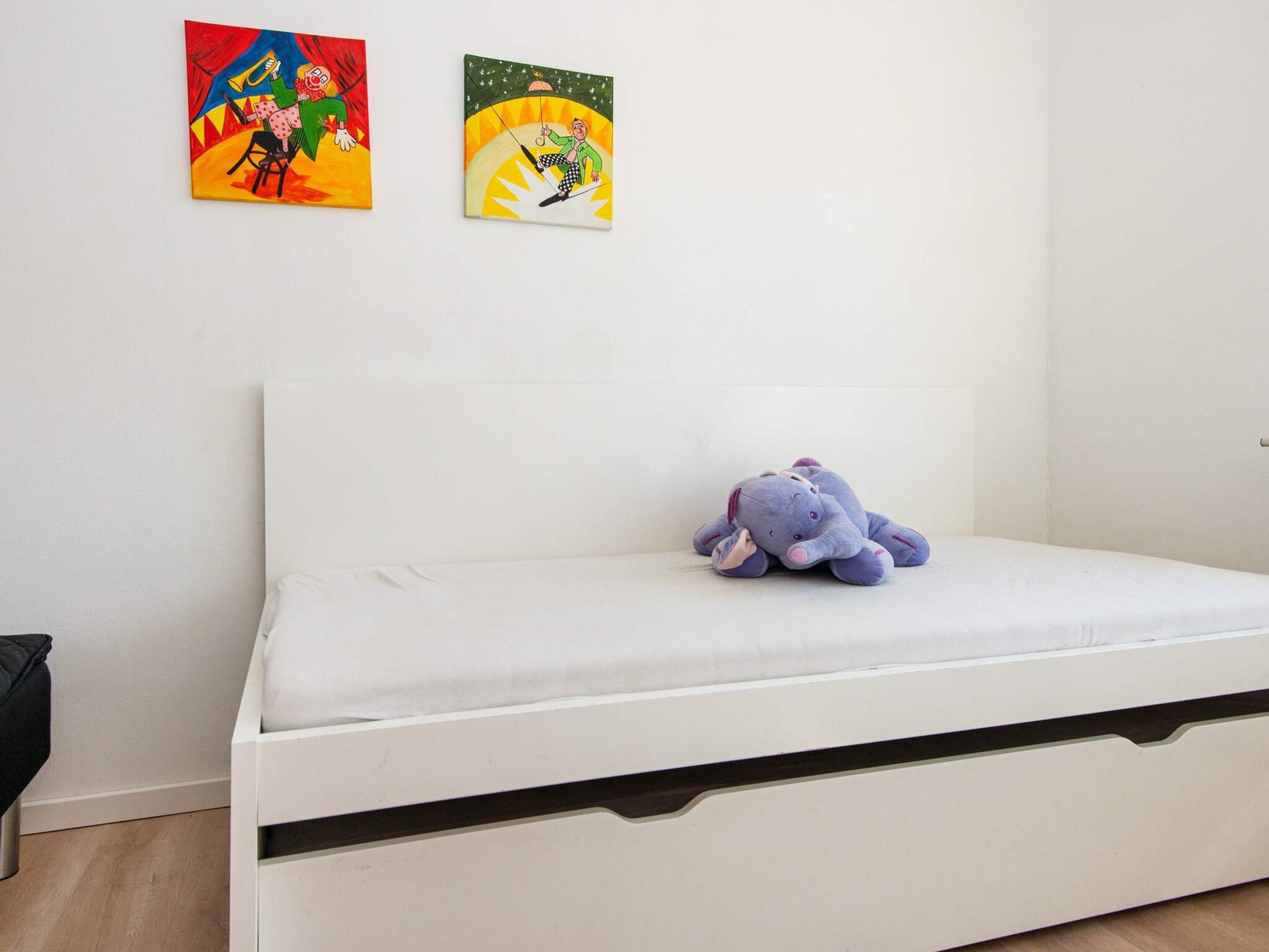 A bedroom with a white bed, a plush toy on the bed, and two colorful paintings on the wall.
