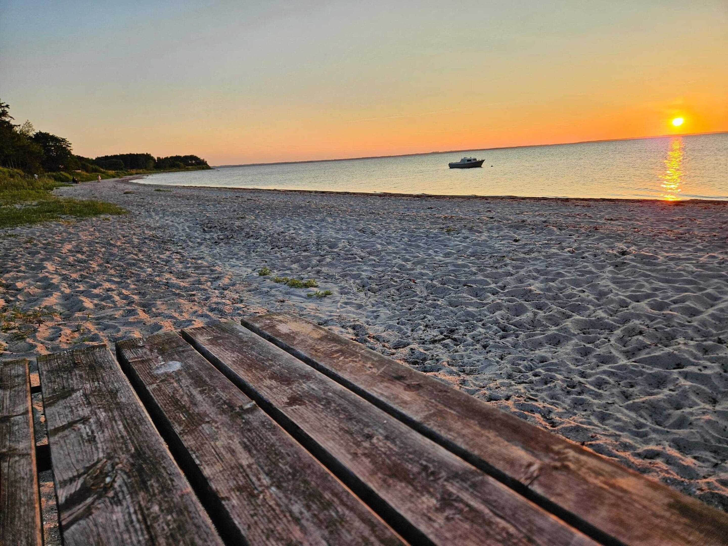A wooden boardwalk leading to a beach with a boat on the water and a sunset in the background.