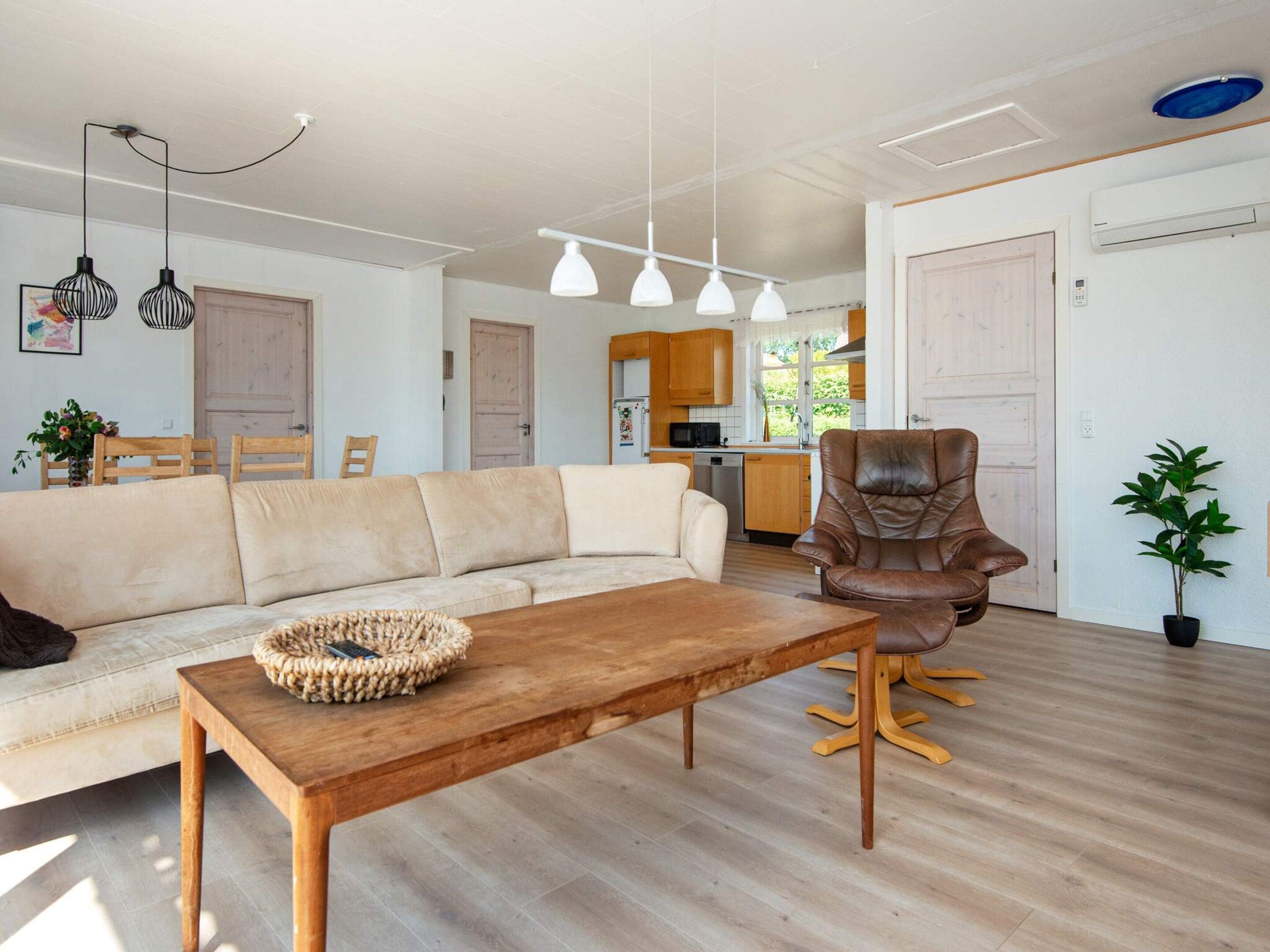A modern living room with a sofa, a wooden coffee table, and a leather armchair. There is a kitchen area in the background with wooden cabinets and a microwave.