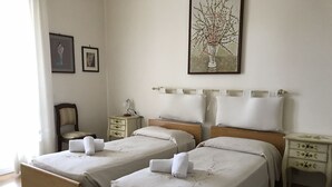 Quarto de hotel com duas camas, uma cadeira, mesa de cabeceira, abajur e um quadro na parede.