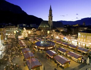 A square with market stalls and a central fountain, surrounded by buildings and a church tower. - Holiday Apartment Krosshof (Nalles)