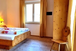 A bedroom with a wooden bed, a nightstand, a lamp, a window with curtains, and a wooden chair. - Holiday Apartment Krosshof (Nalles)