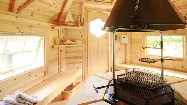 Interior de cabana com teto de madeira, churrasqueira, janela e prateleira com artigos de higiene pessoal.